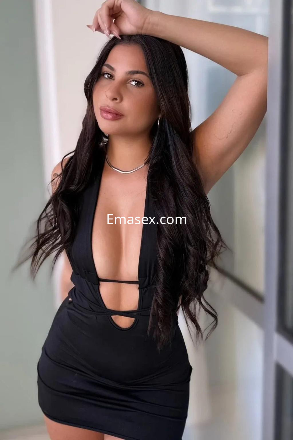 Escort en Valencia con vestido negro ajustado, escote pronunciado y cabello largo y oscuro, posando con una mano en la cabeza.