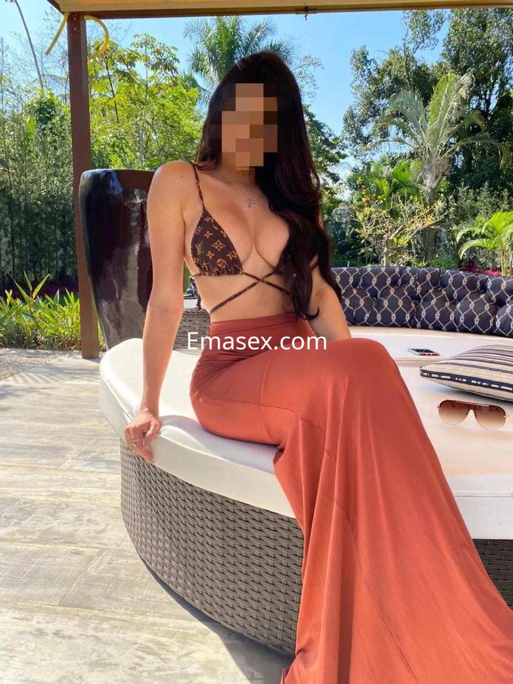 Escort en Valencia sentada en un sofá exterior, con un top de bikini marrón y una falda larga color terracota.