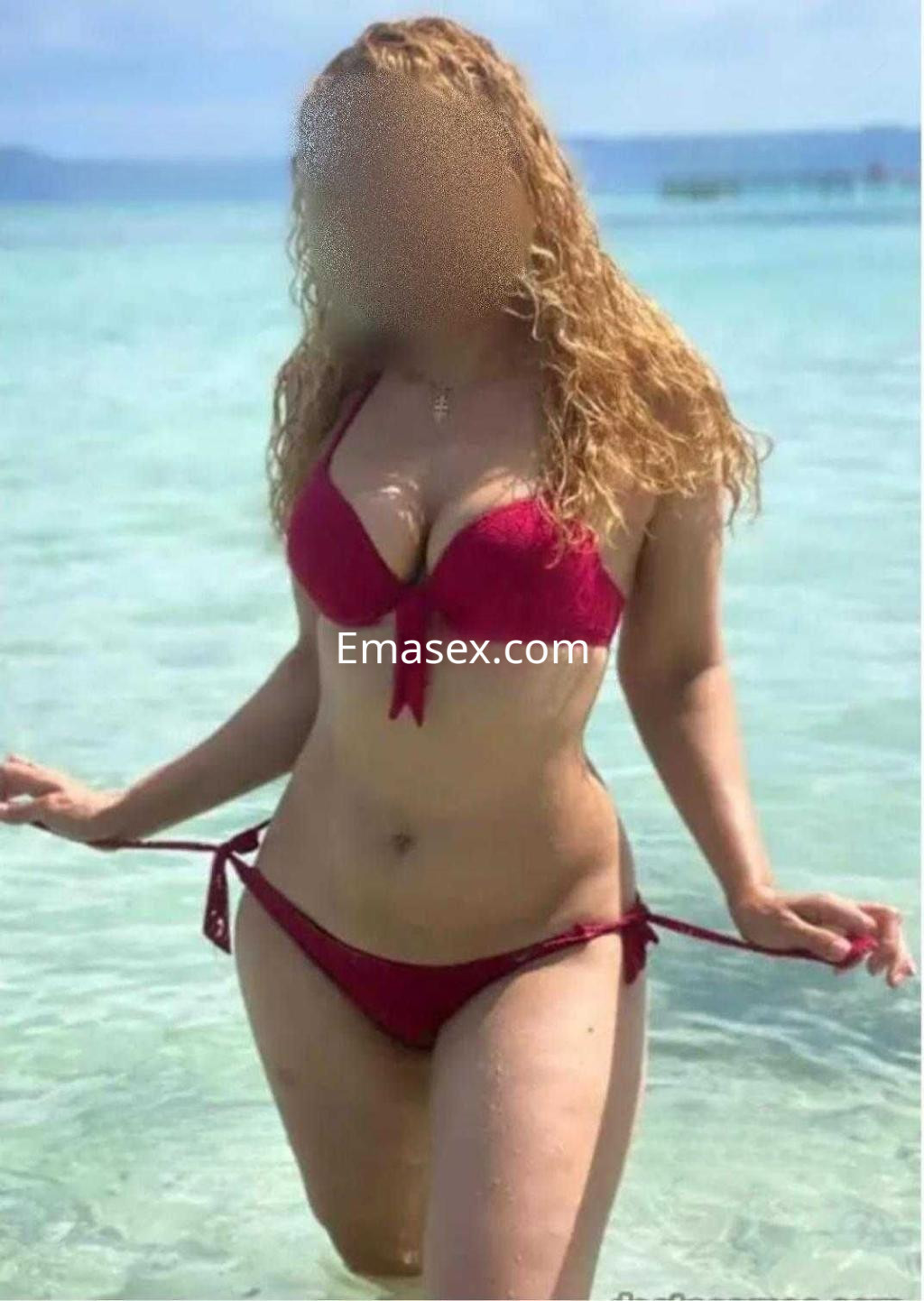 Escort en Zaragoza con cabello rizado y rubio, luciendo un bikini rojo en una playa con agua clara.
