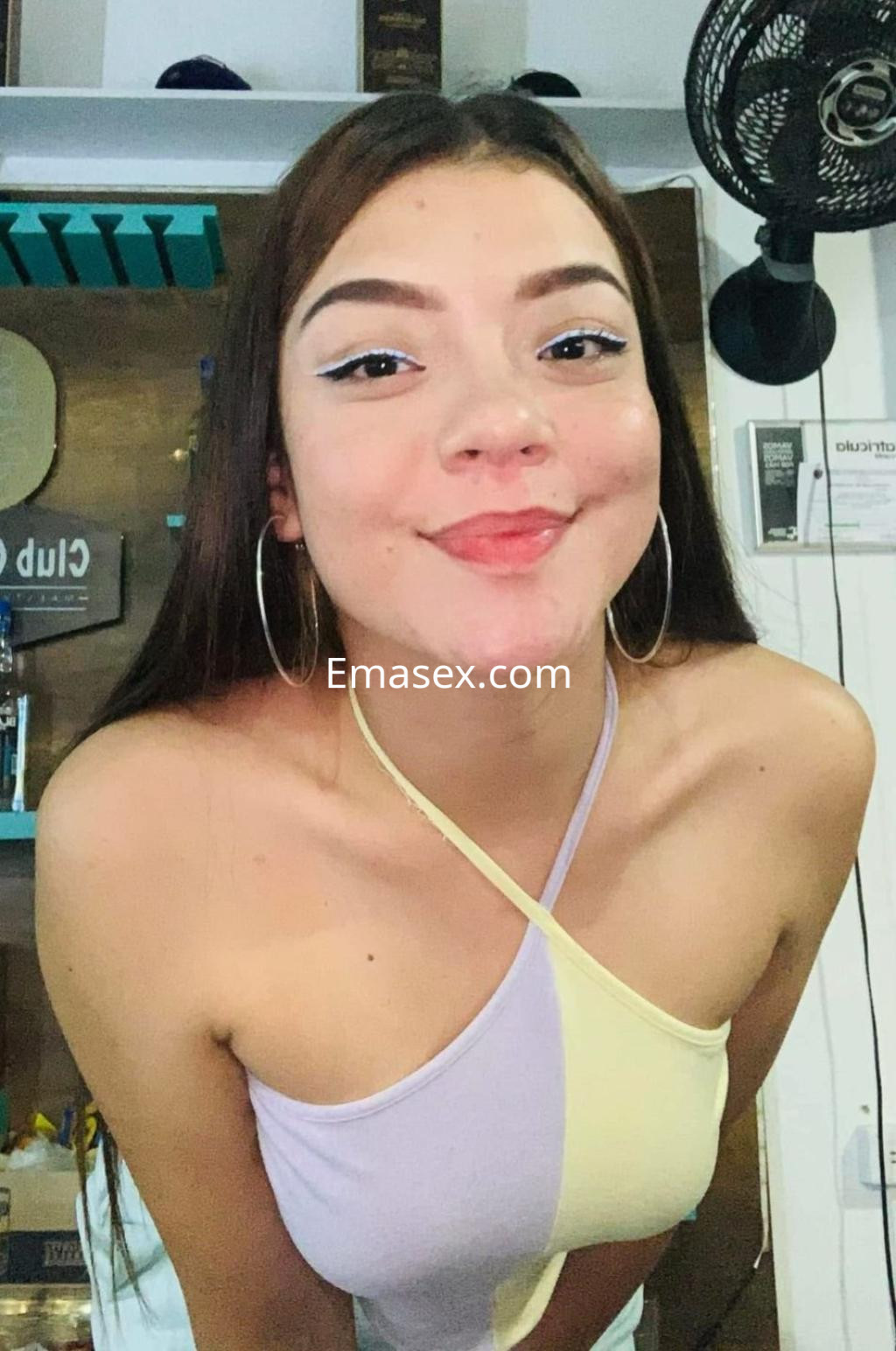 Escort en Sevilla sonríe mientras viste una blusa de tirantes en tonos pastel y aretes grandes, en un ambiente acogedor.