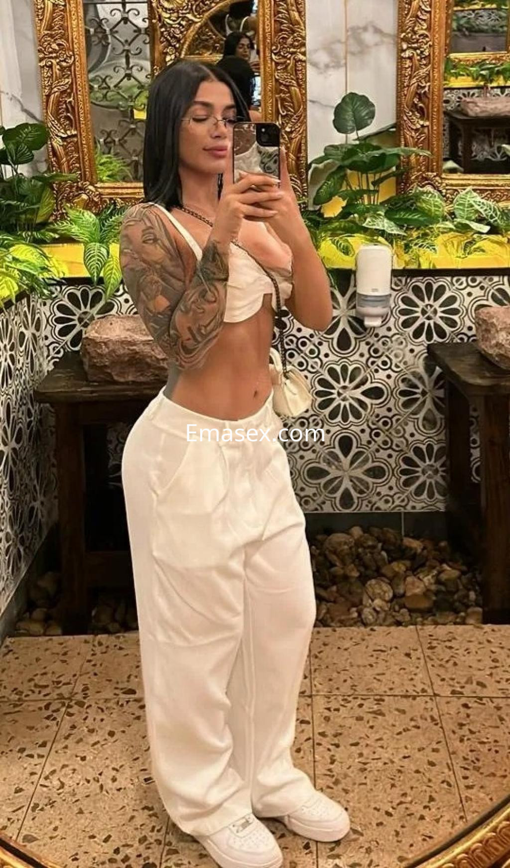 Escort en Zaragoza con top claro y pantalones blancos, posando frente a un espejo con detalles decorativos.