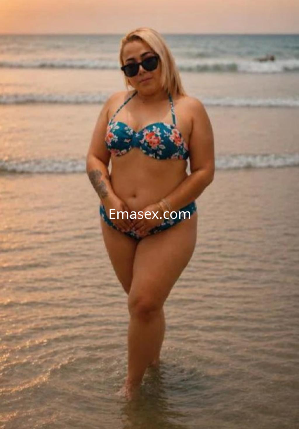 Escort en Cádiz, posando en la playa con un bikini floral, gafas de sol y el mar de fondo al atardecer.