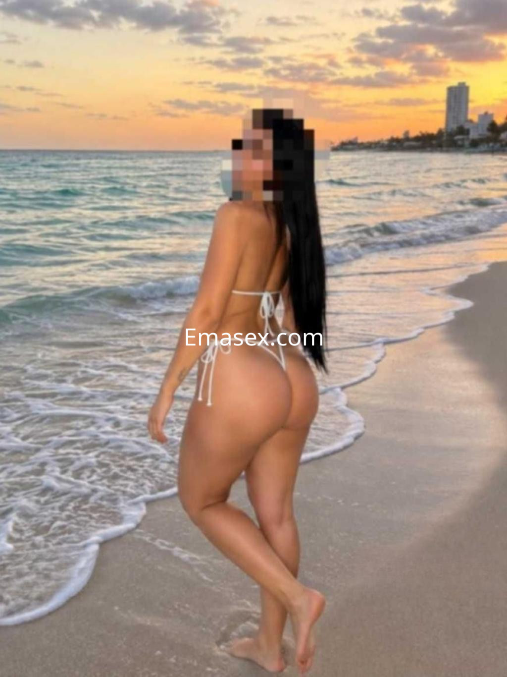Escort de cabello largo y lacio, luciendo un bikini blanco, posando de espaldas en la playa al atardecer en Alicante.