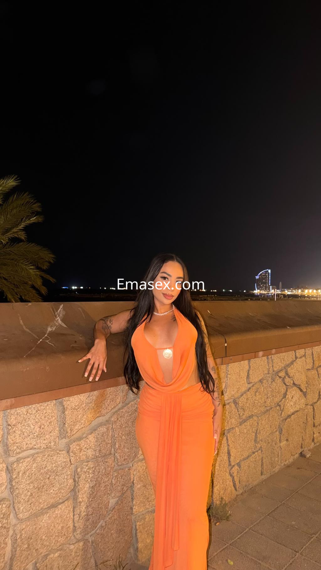 Escort de Pamplona con cabello largo y oscuro, vistiendo un vestido naranja, posando junto a un muro en la noche.