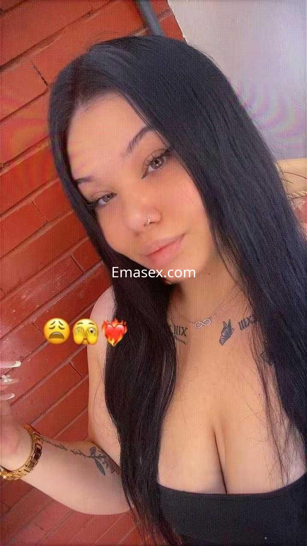 Escort en Almería con cabello largo y lacio, posando frente a una pared de ladrillo, mostrando un escote y tatuajes.