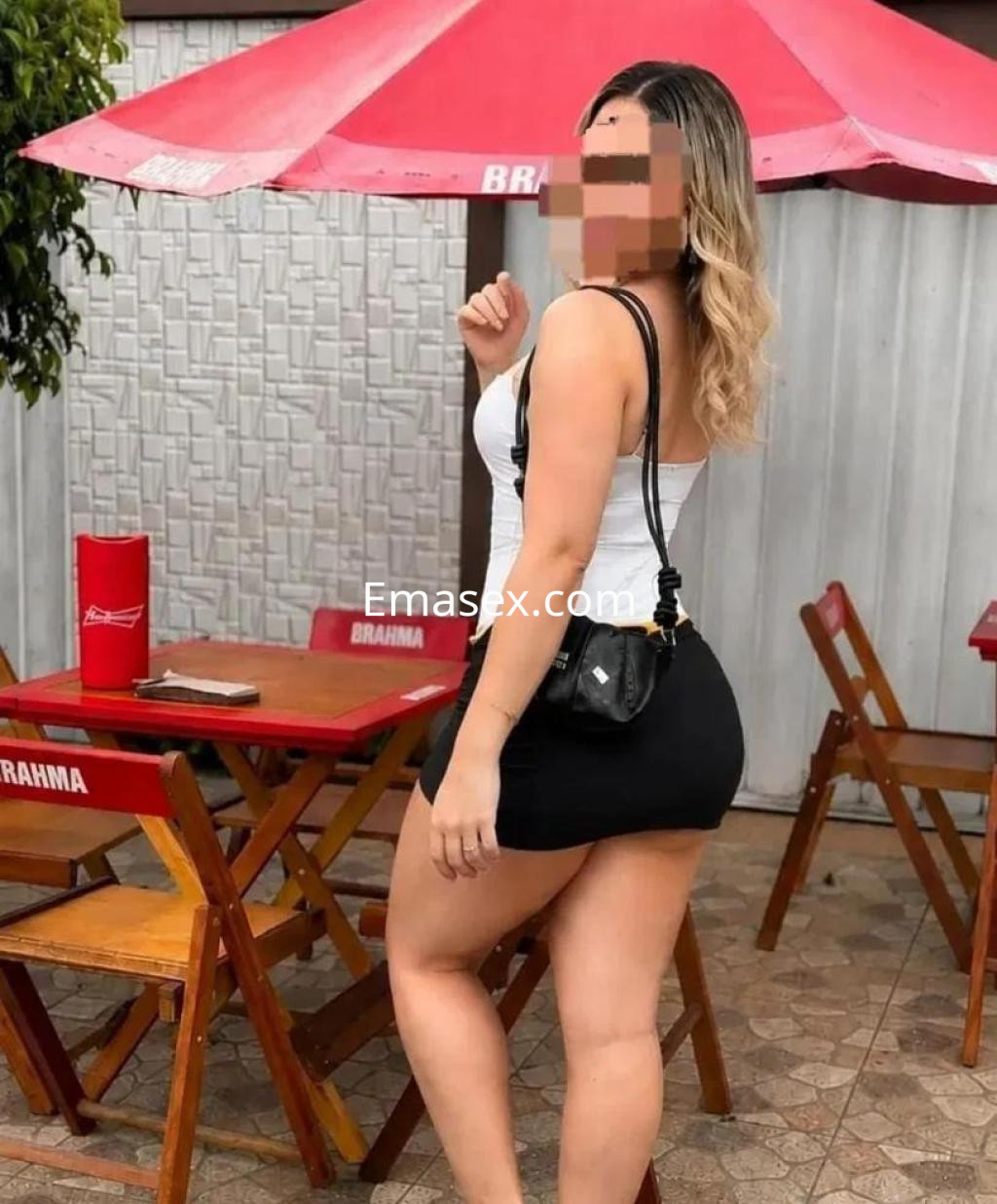 Puta de Almería viste una blusa blanca ajustada y una falda corta negra, posando con un bolso en un entorno al aire libre.