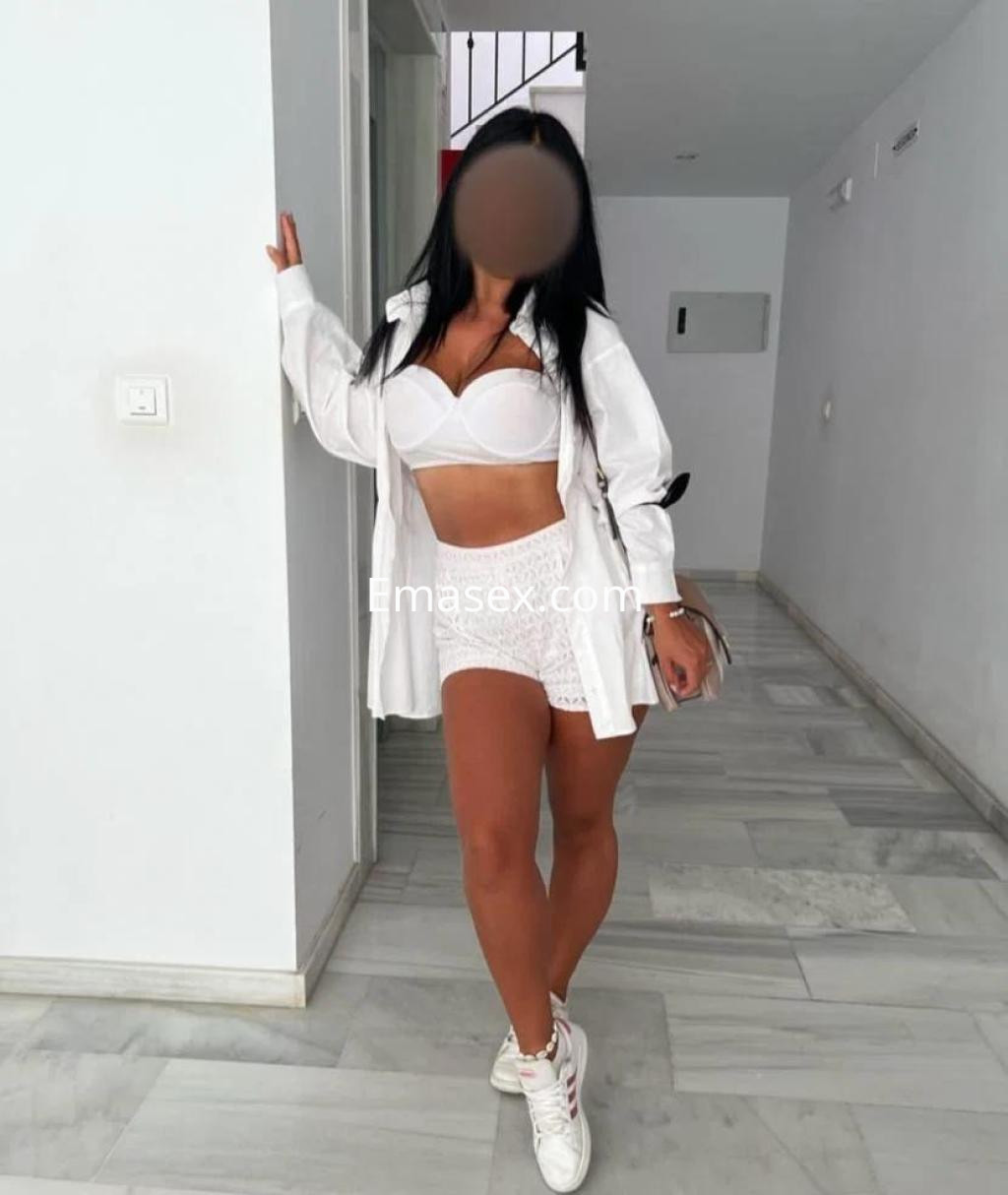 Escort en Coruña con blusa blanca y shorts, posando de forma relajada en un ambiente interior claro.