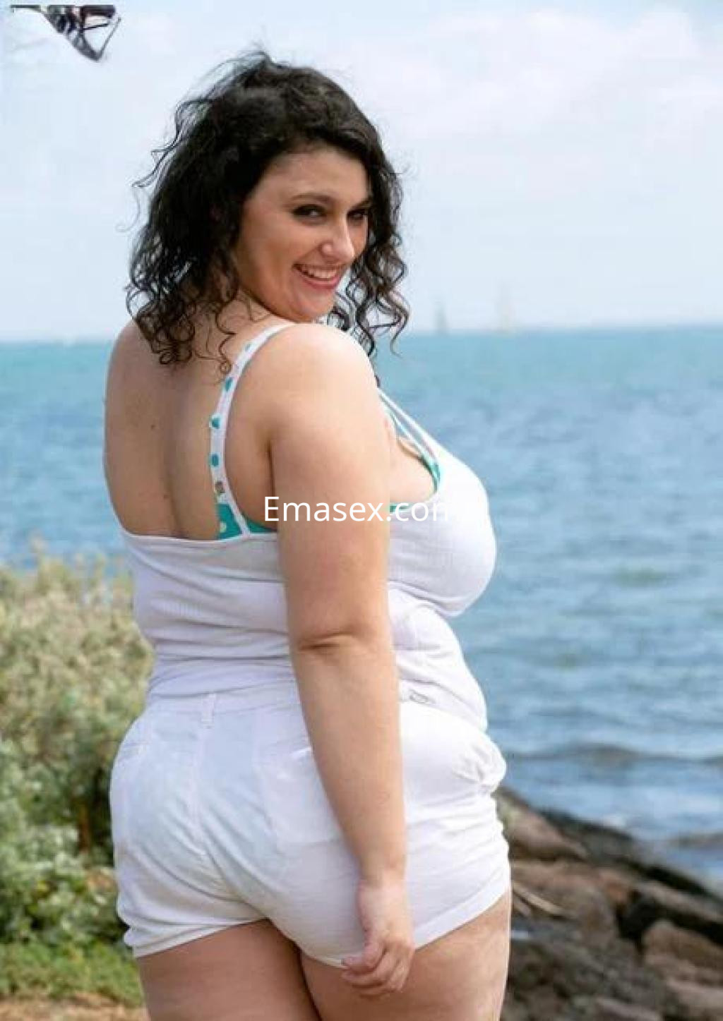 Escort posando de espaldas junto al mar en Oviedo, viste un top blanco y shorts cortos, con cabello rizado.
