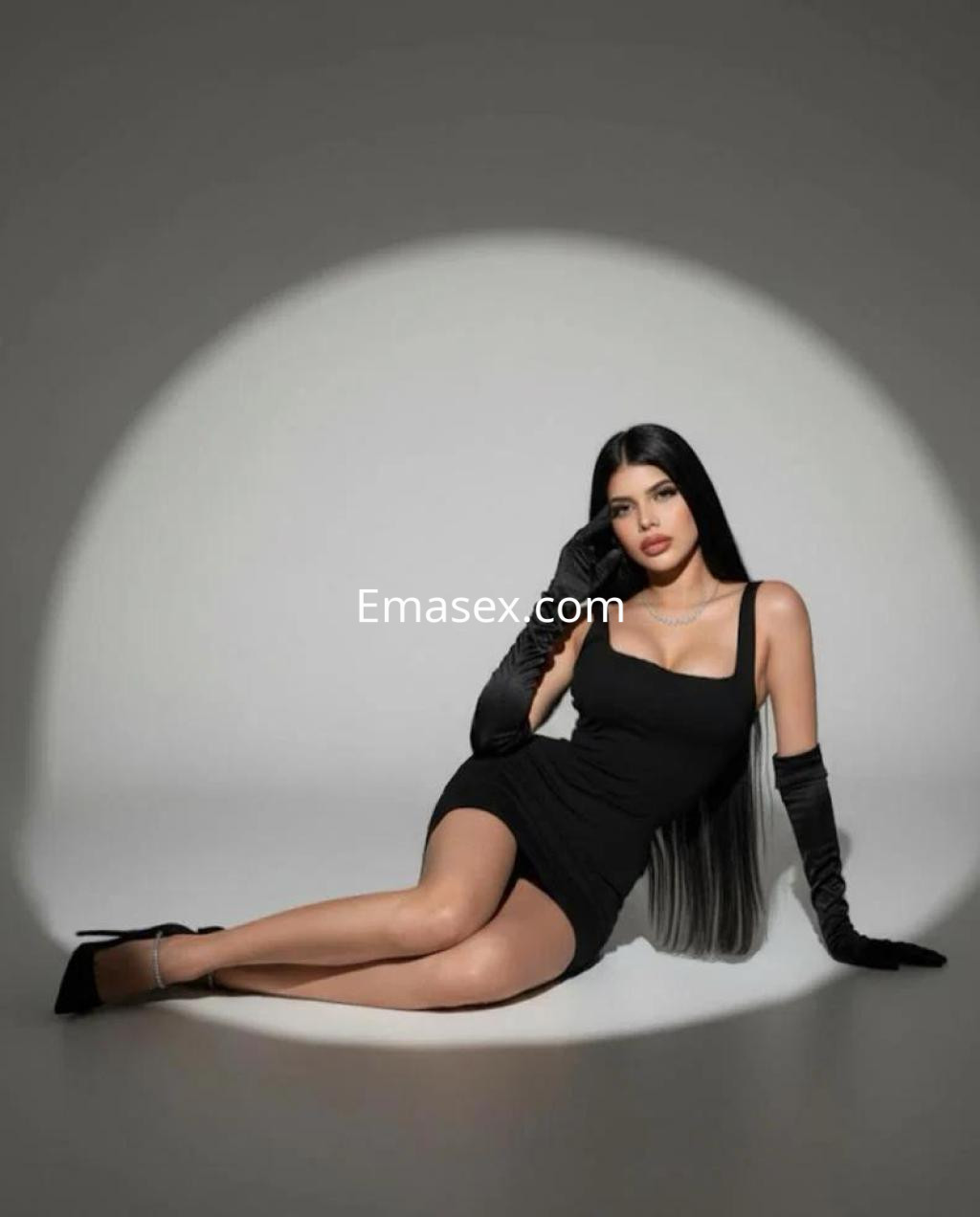 Escort en Las Palmas, posando con un vestido negro ajustado y guantes largos, sobre un fondo iluminado.