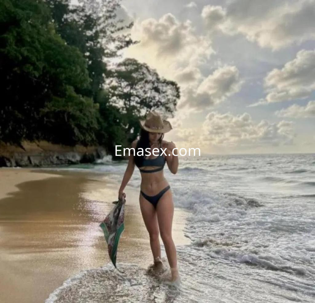 Escort en Salamanca, de pie en la orilla del mar con un bikini negro y sombrero, sosteniendo una toalla.