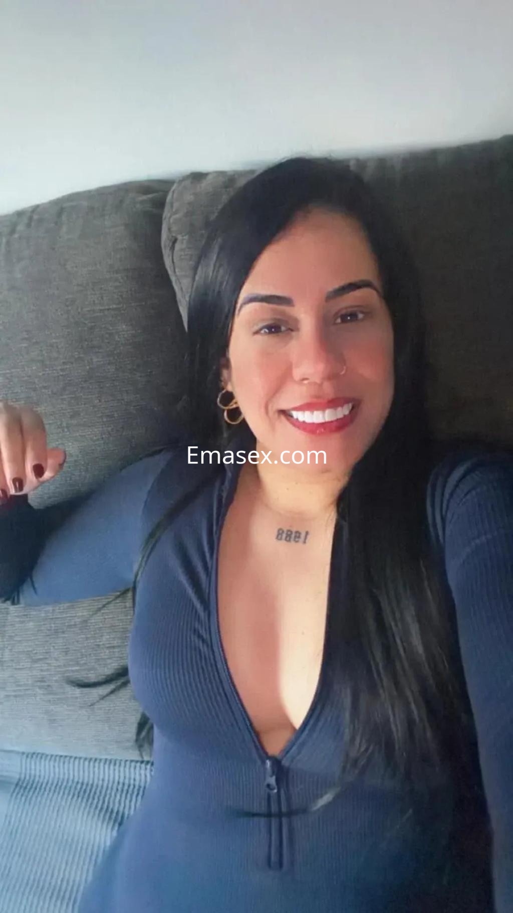 La escort en Toledo sonríe mientras se relaja en un sofá, luciendo un vestido azul con un escote pronunciado.
