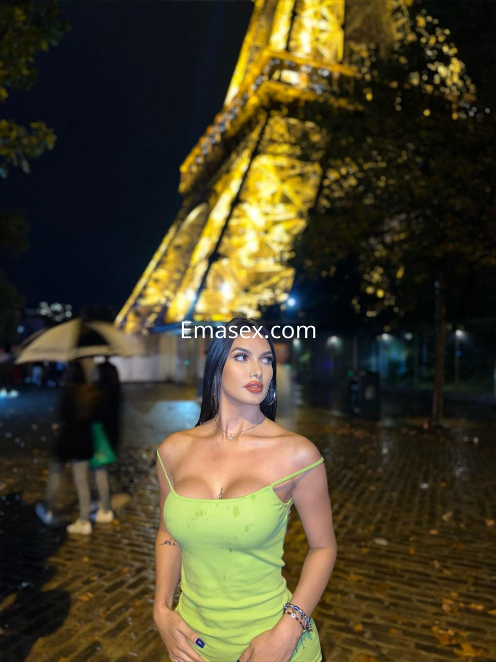 La escort en Toledo viste una blusa verde ajustada, posando frente a la Torre Eiffel iluminada por la noche.