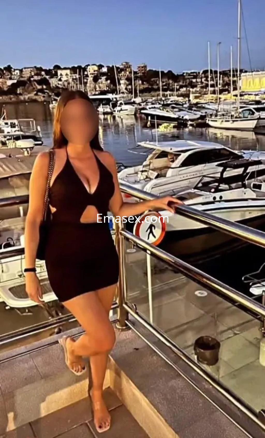 La escort en Valencia luce un vestido negro ajustado y posa junto a un puerto lleno de yates al atardecer.