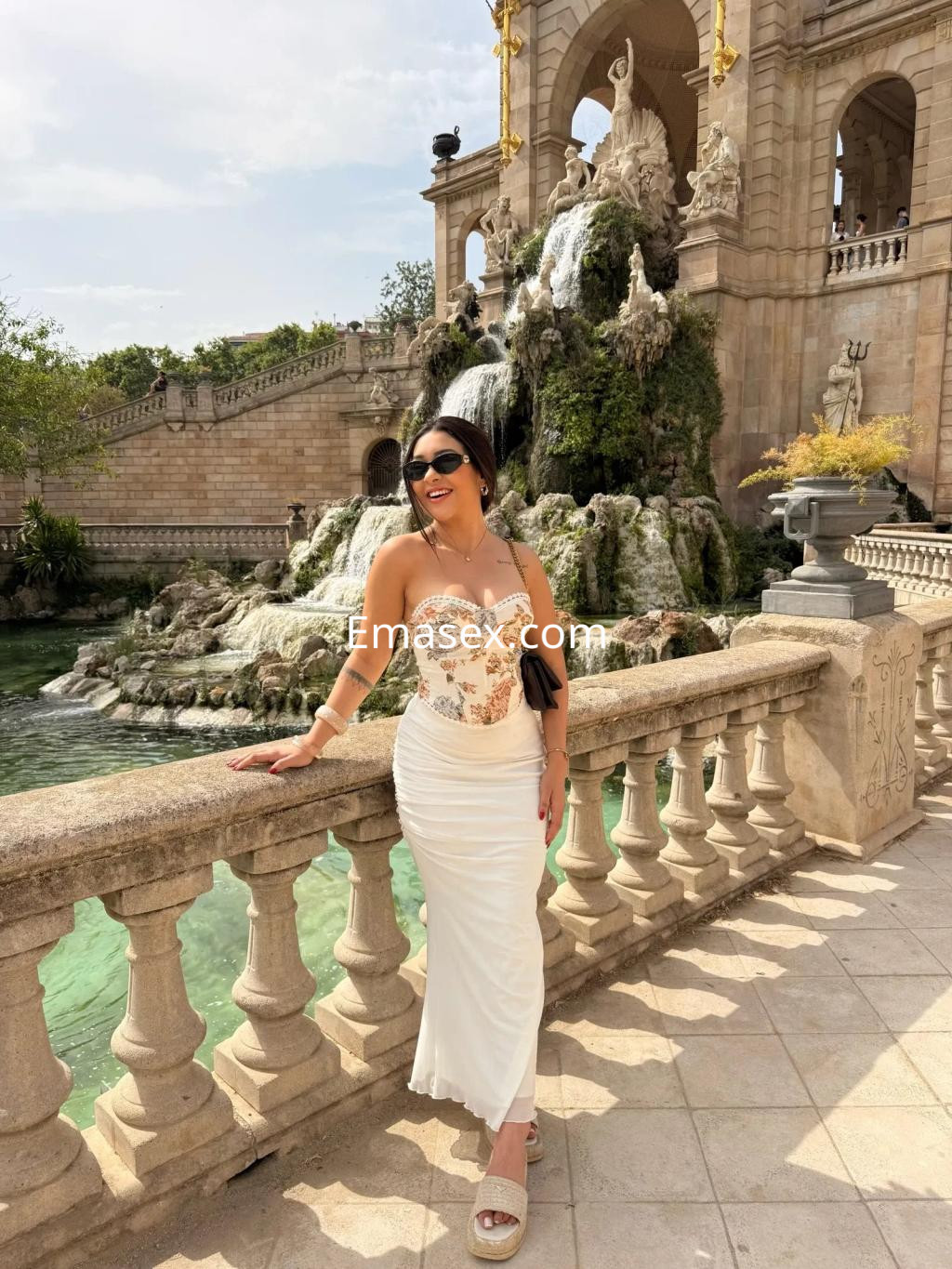 Puta posando con un vestido blanco ajustado en un hermoso escenario de Valencia, junto a una fuente y vegetación.