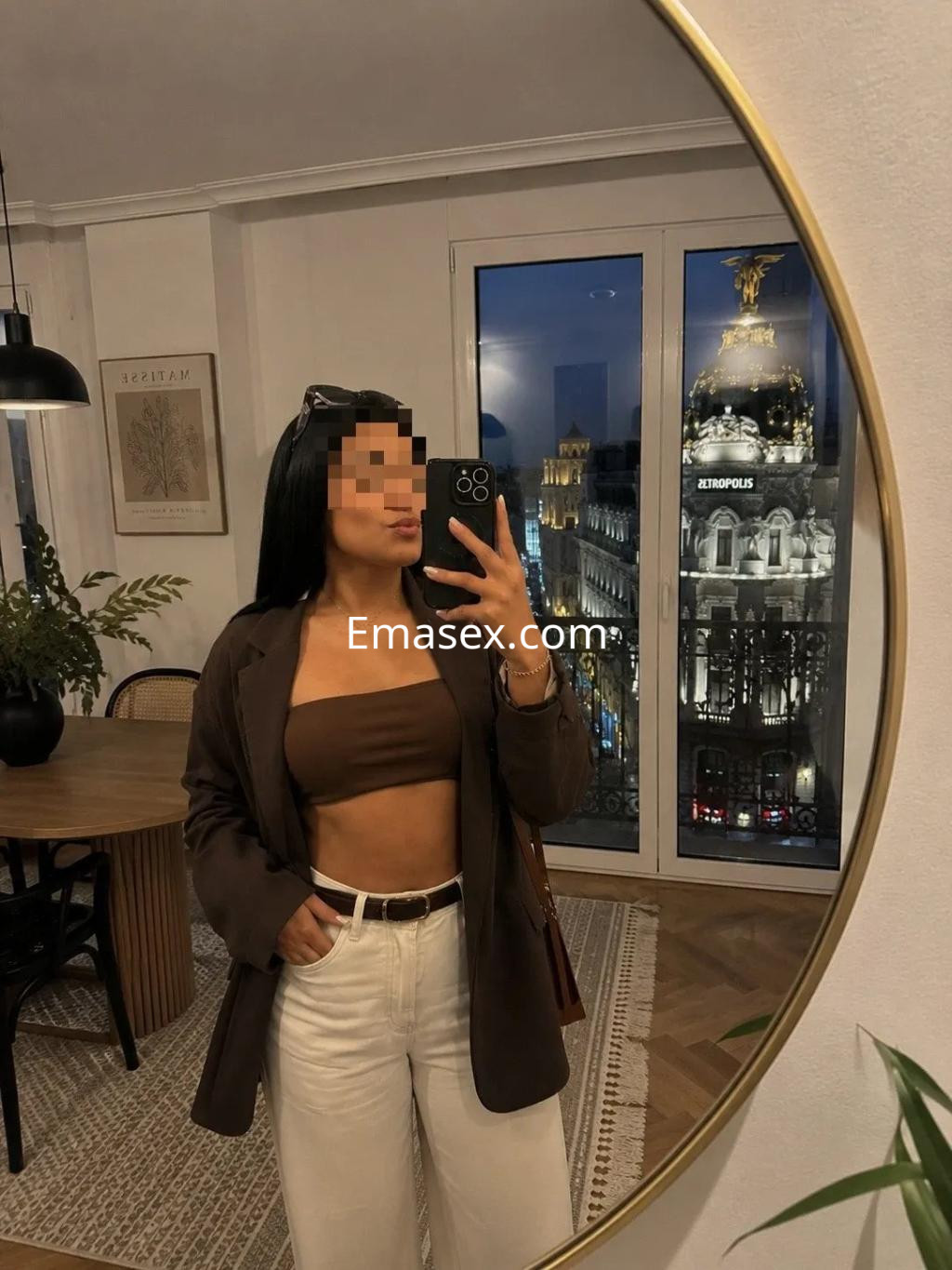 Escort en Valladolid, posando en un espejo con blusa marrón y pantalones claros, ante un fondo urbano nocturno.