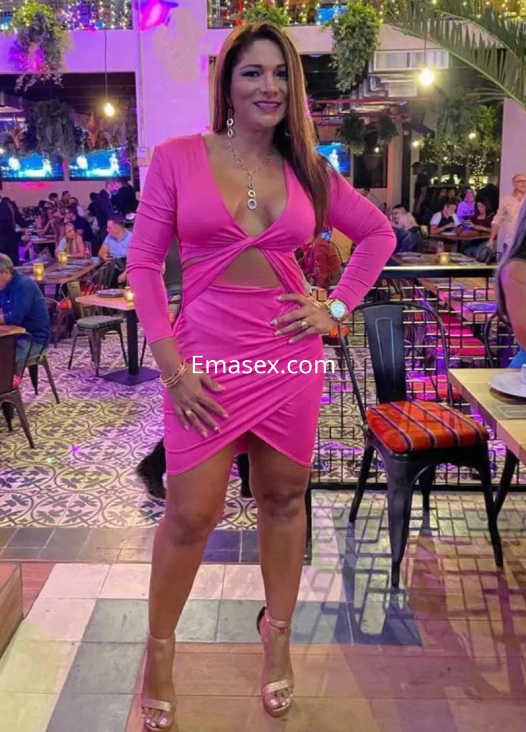 Puta en Sevilla con vestido rosa ajustado y escote, posa en un bar con ambiente alegre y luces.