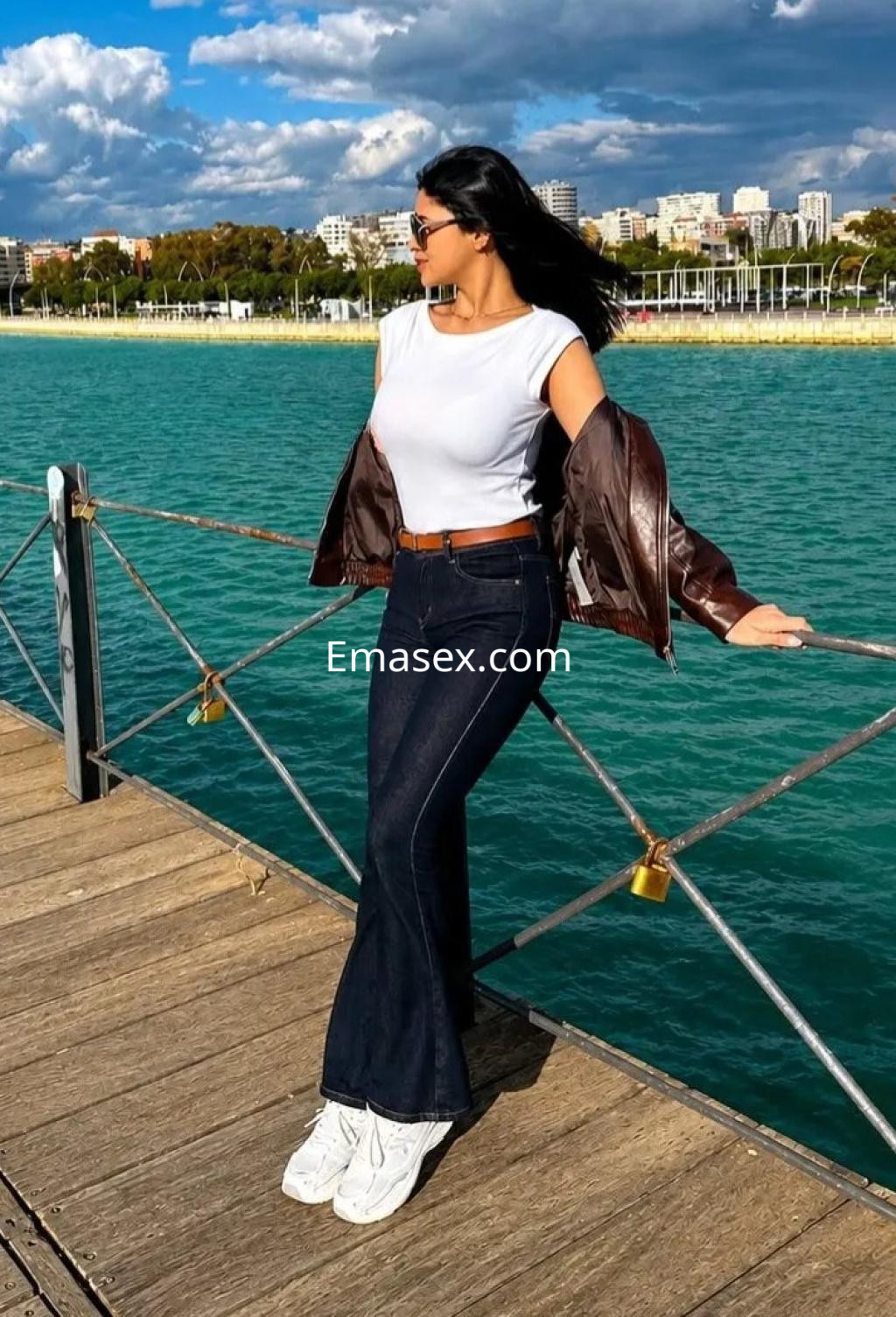 Escort en Málaga, posando junto al mar con una camiseta blanca, chaqueta marrón, jeans oscuros y zapatillas.