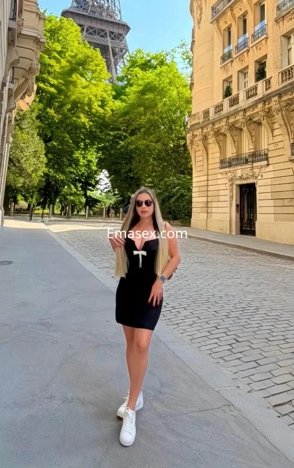 Escort en Palma de Mallorca luce un vestido negro ajustado, con gafas de sol y zapatillas, caminando por una calle arbolada.