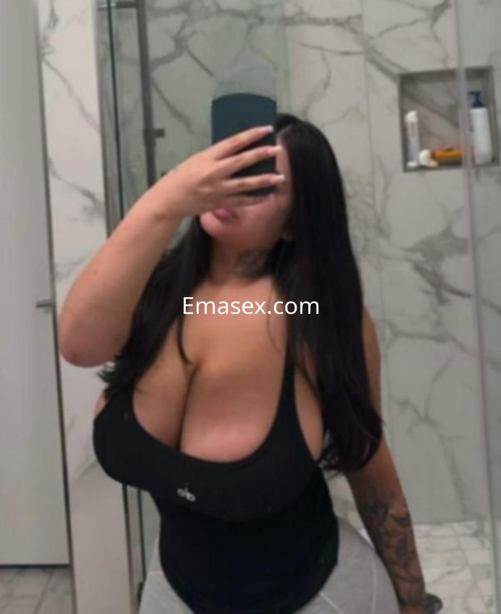 Escort en Vigo con cabello largo y oscuro, luciendo una camiseta negra y pantalones ajustados en un baño moderno.