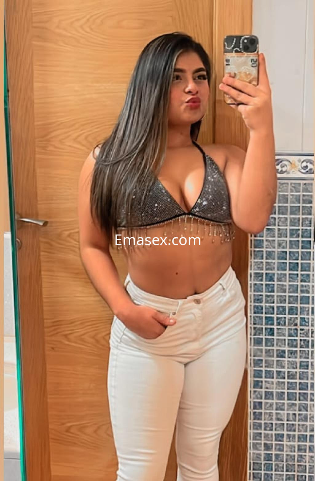 Puta en Valencia con top brillante y casi sin mangas, posando frente al espejo con pantalones blancos.