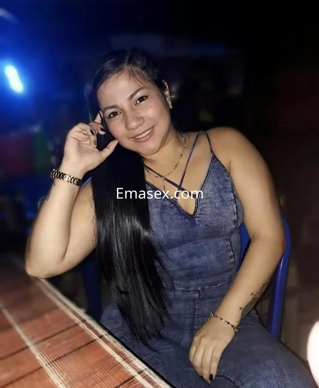Puta en Burgos sentada en una mesa, luciendo un vestido de mezclilla y sonriendo con el cabello largo y lacio.