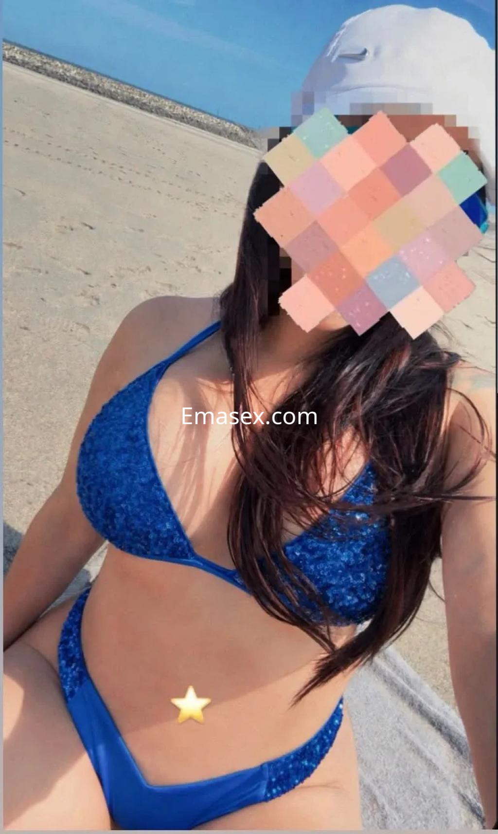 Escort posando en la playa de Coruña, con un bikini azul brillante y gorra blanca, mostrando su cabello largo.