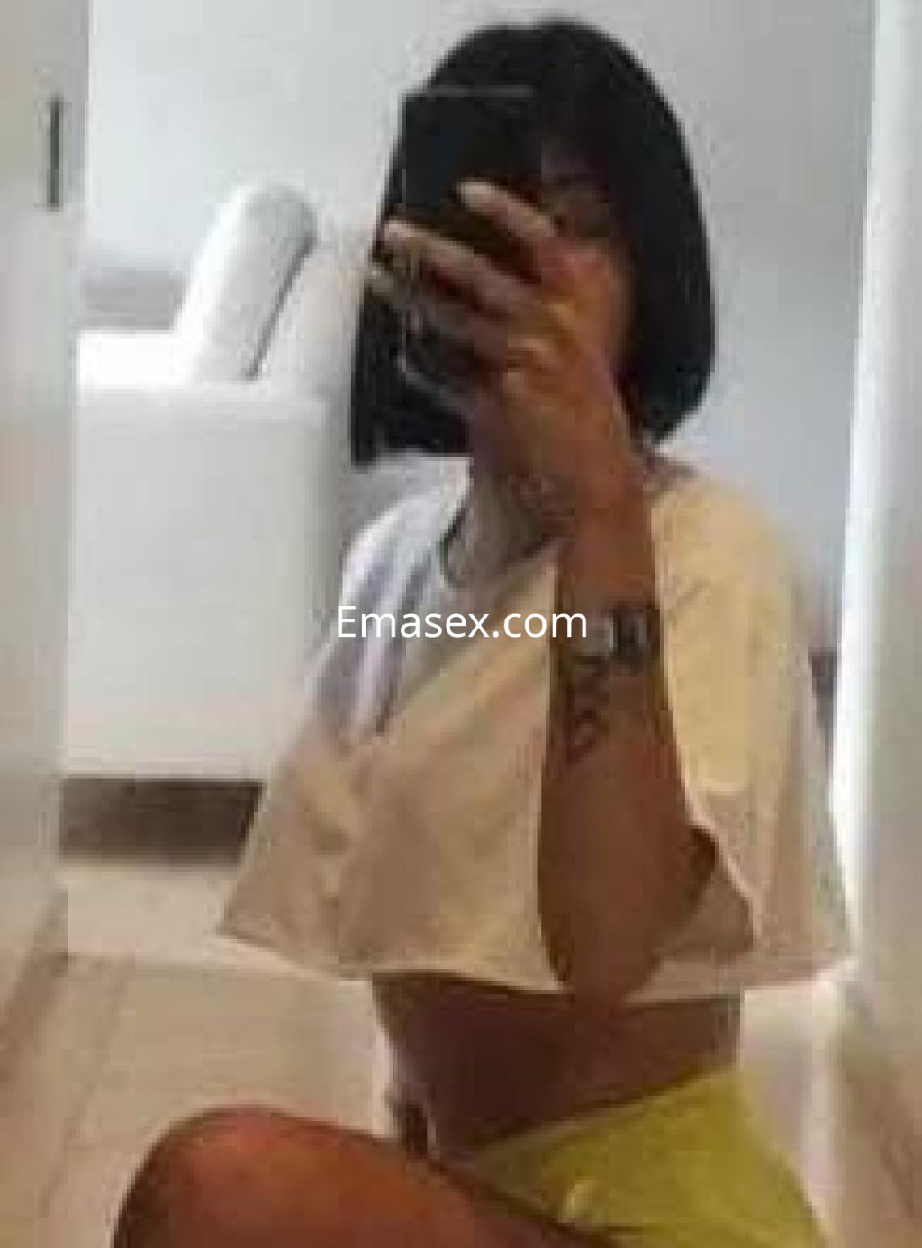 Escort en Girona, con cabello corto y oscuro, viste una blusa corta blanca y una falda amarilla, posando en un espejo.