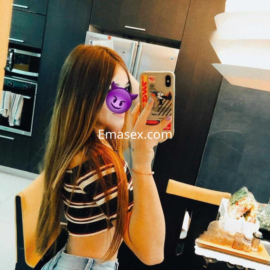 Escort en Granada con cabello largo y lacio, vistiendo una blusa de rayas y tomando una selfie en la cocina.