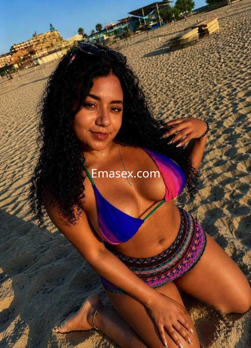 Puta de Huelva se encuentra en la playa, luciendo un bikini multicolor y shorts cortos, posando sonriente.