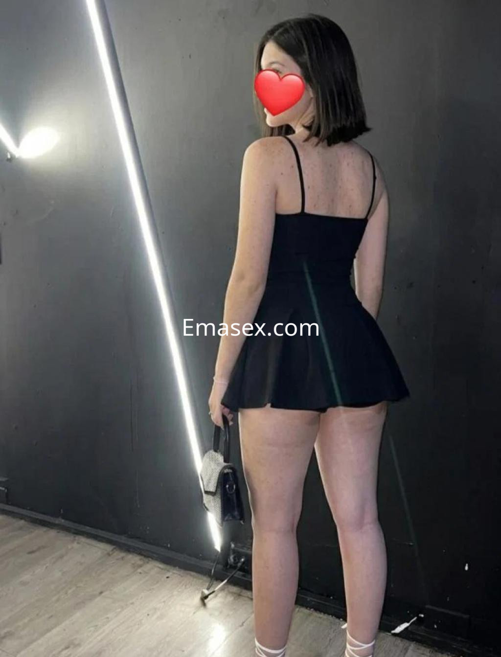 Escort de Toledo con vestido negro corto y tirantes, posando de espaldas en un ambiente moderno.