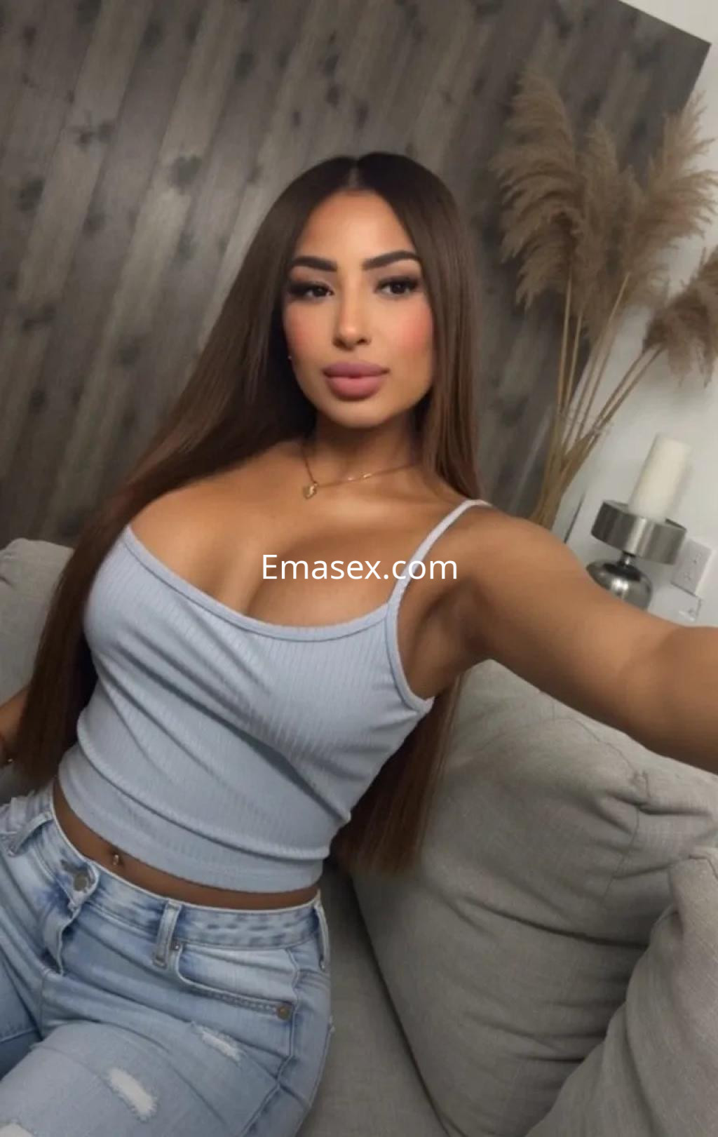 Escort en Valencia con cabello largo y lacio, vistiendo blusa sin mangas azul y jeans ajustados, sonríe en un ambiente acogedor.