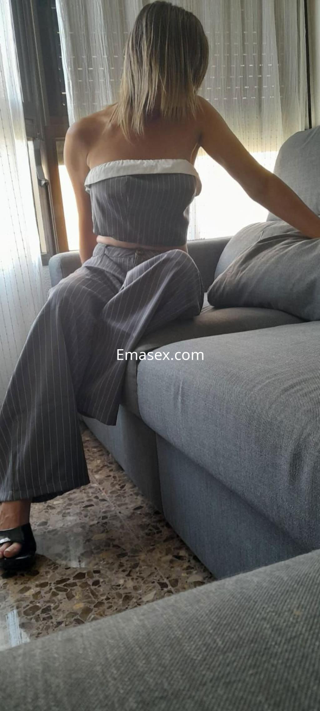 Escort en Valencia sentada en un sofá, lleva un top sin mangas y pantalones a rayas grises, con cabello rubio.