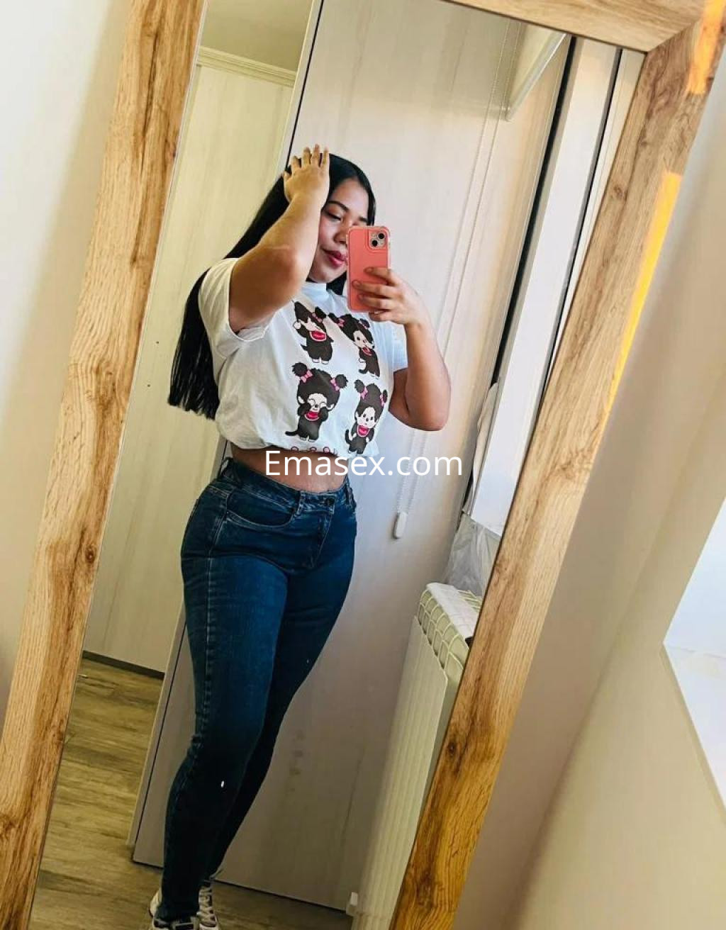 Escort en Zamora posando frente a un espejo, viste una camiseta blanca y jeans ajustados, con cabello largo y lacio.