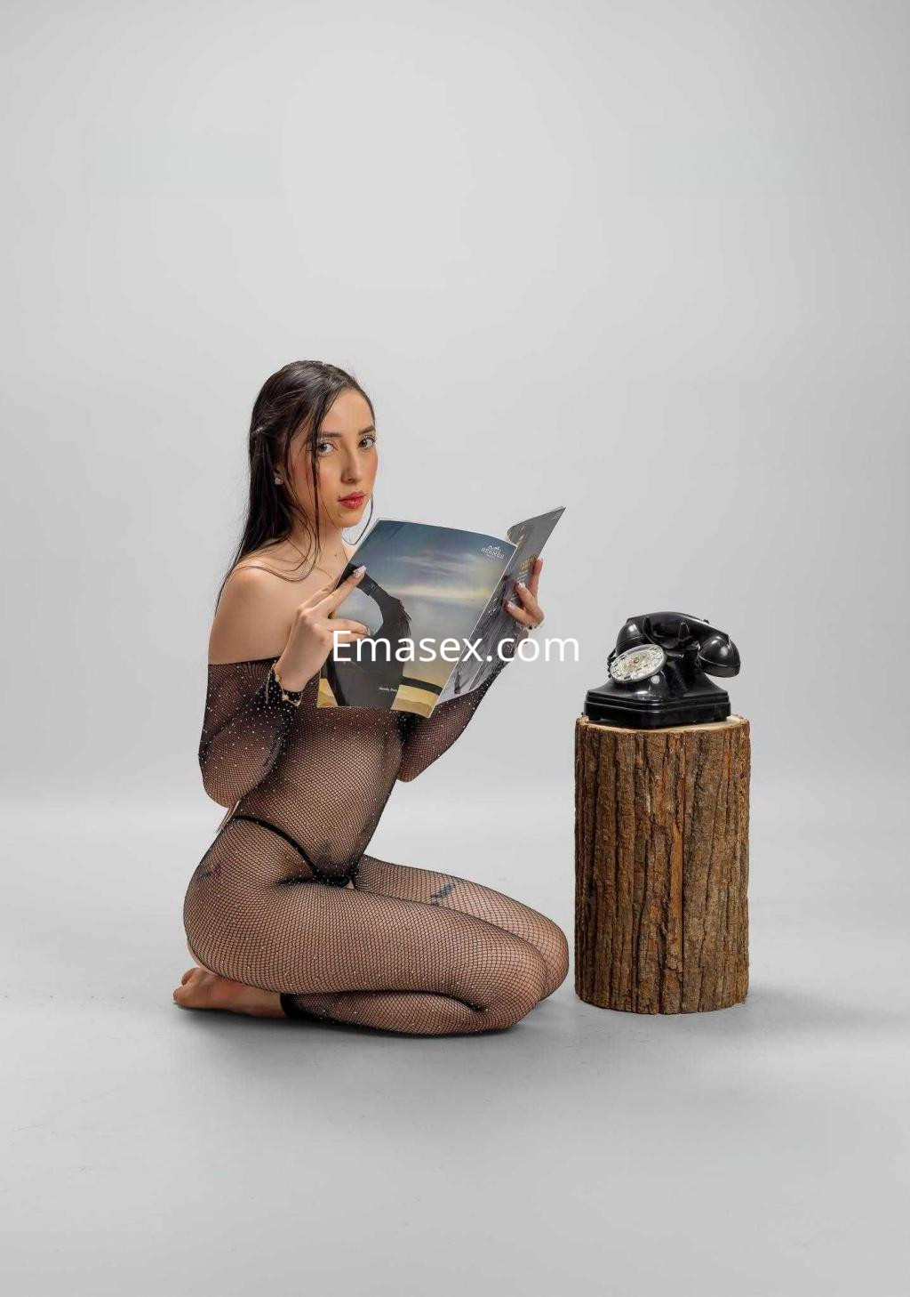 Escort en Zaragoza luce un traje de malla transparente mientras sostiene una revista, sentada sobre las rodillas.