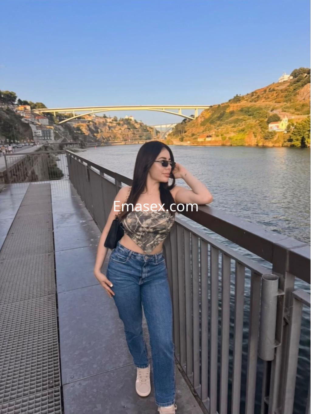 Escort en Vigo con top de estampado camuflaje y jeans ajustados, apoyada en una barandilla junto al agua.