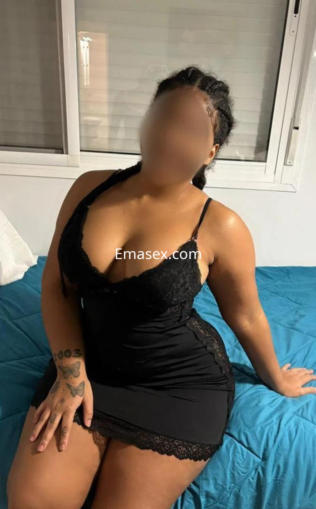 Escort en Sevilla, sentada sobre una cama con sábanas azules, vistiendo un vestido negro de encaje.