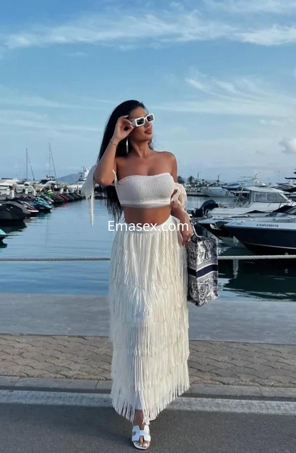 Puta en Palma de Mallorca, luciendo un top blanco y una falda larga con flecos, posando junto al puerto.
