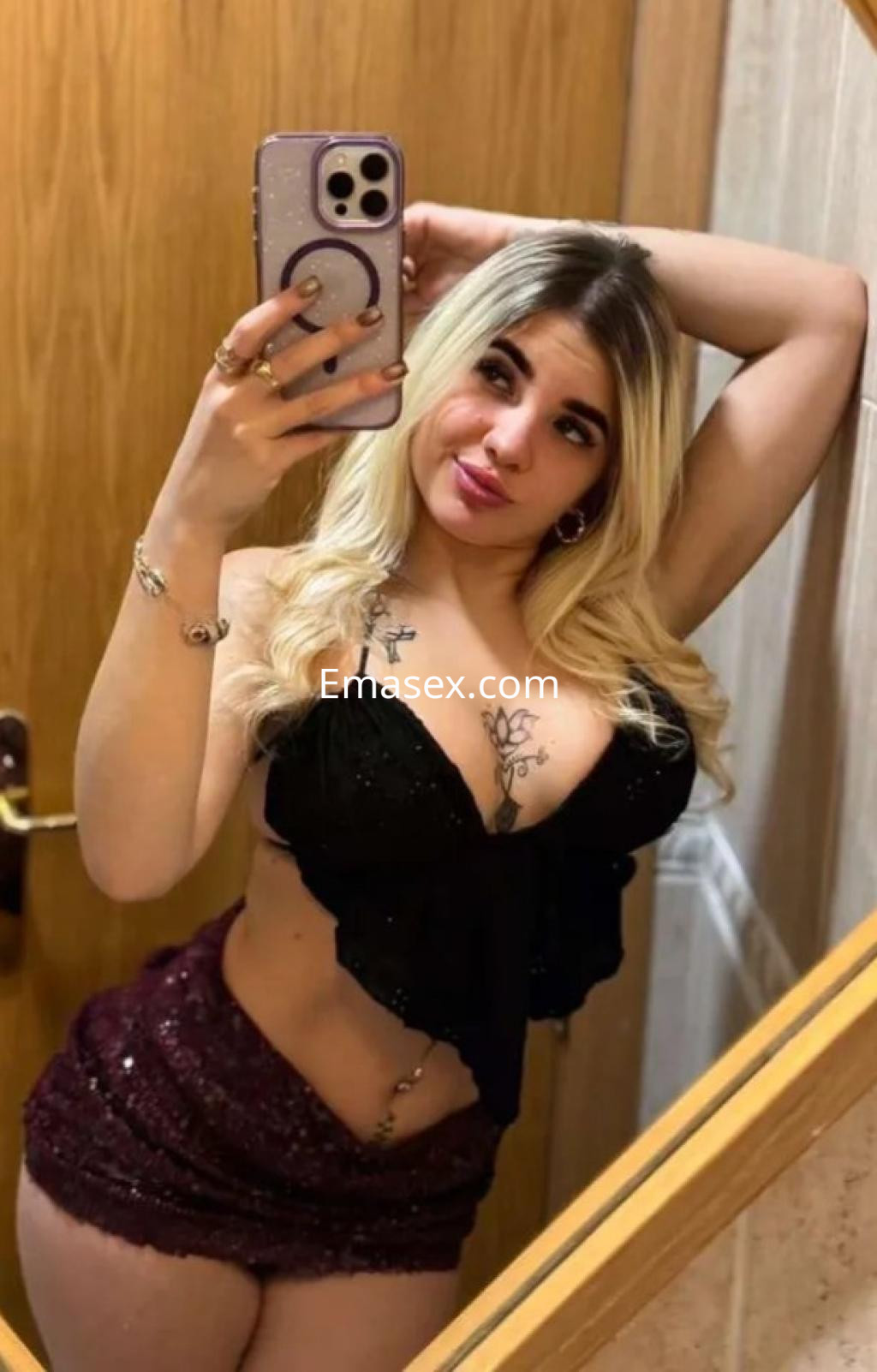 Escort en Málaga con cabello rubio, blusa negra y falda corta, posando frente a un espejo.