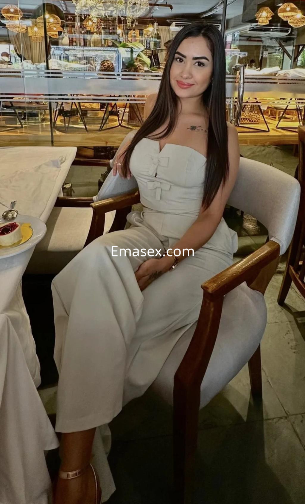 Puta sentada en una silla en un restaurante de Málaga, luciendo un conjunto blanco sin mangas y cabello largo.