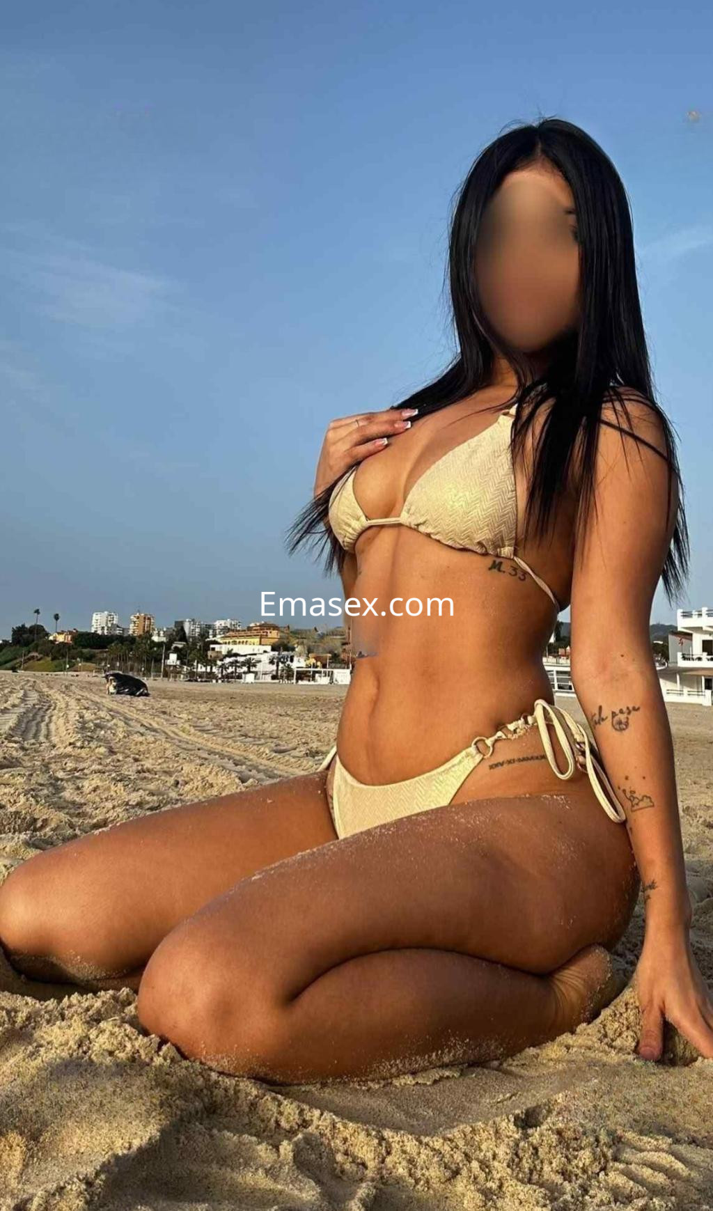 Puta de piel bronceada, sentada sobre la arena en la playa de Cádiz, luciendo un bikini dorado y tatuajes.