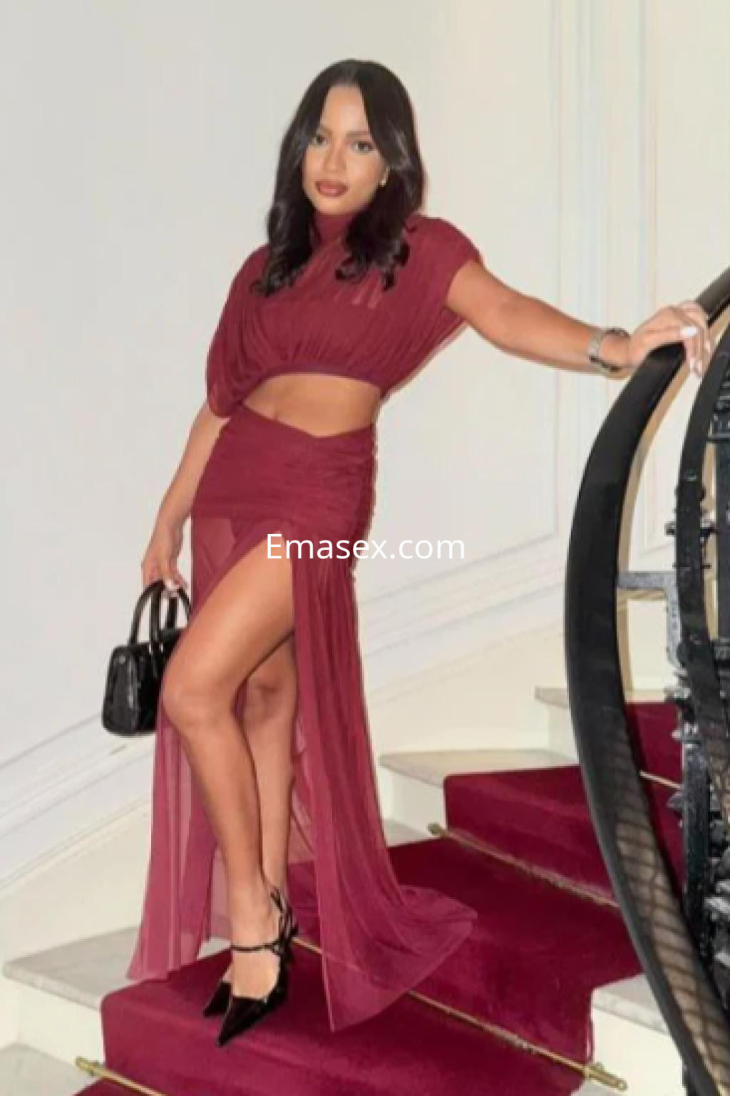 Escort con cabello oscuro y ondulado, viste un vestido burdeos con abertura lateral, posando en una escalera en Santander.