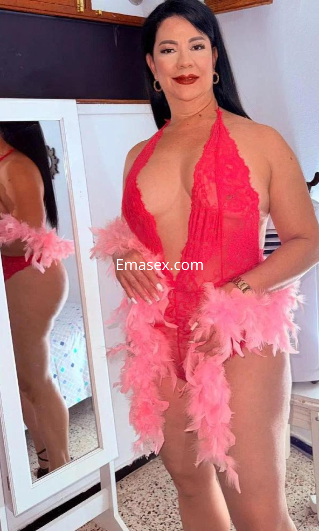 Escort en Las Palmas, posando en un vestido de encaje rojo con plumas rosas, frente a un espejo.