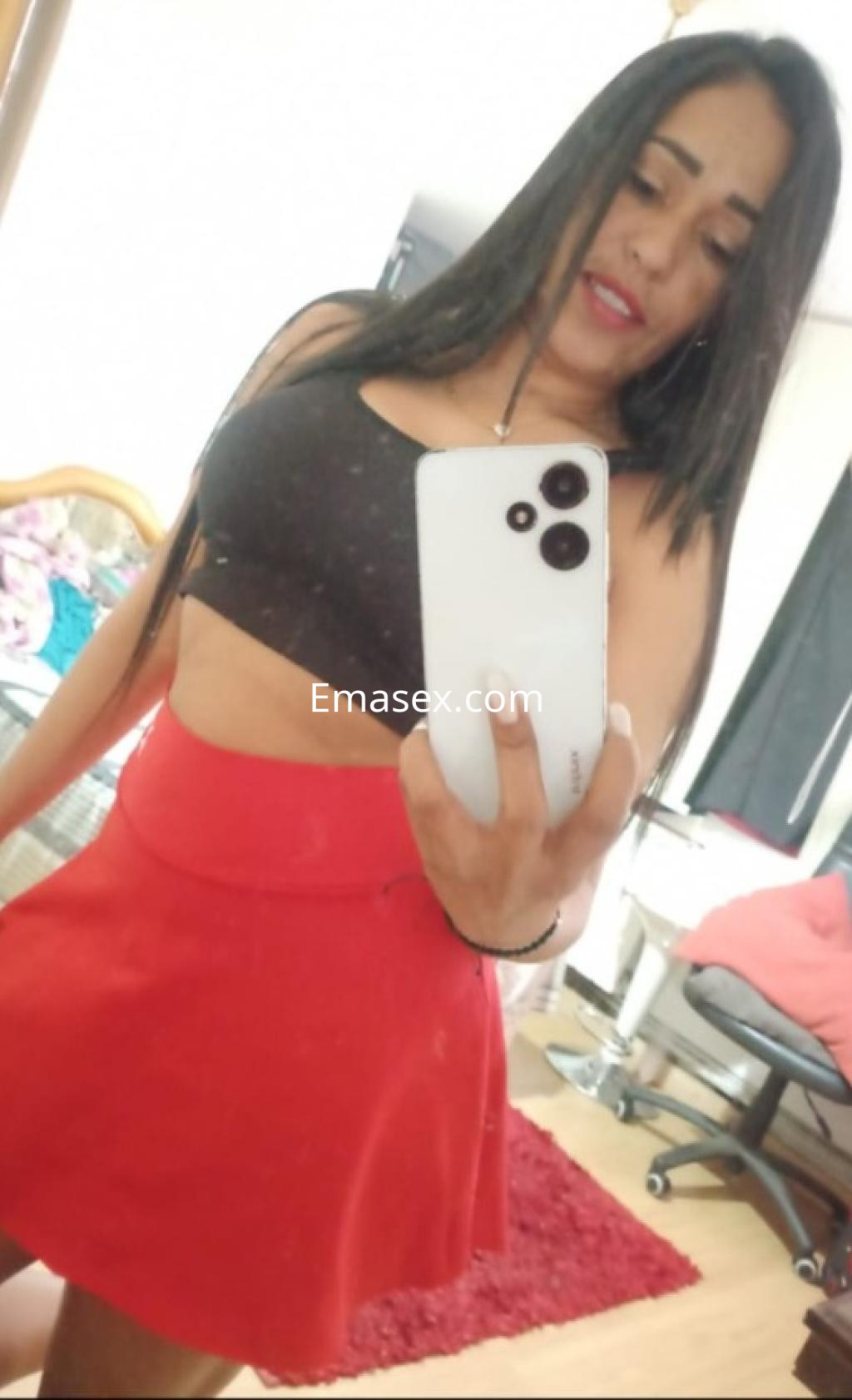 Puta en Pontevedra con cabello largo y lacio, viste un top negro y una falda roja, sonriendo ante el espejo.