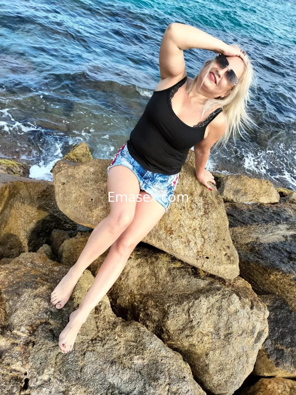 Escort en Albacete sentada sobre rocas junto al mar, luciendo camiseta negra y shorts de mezclilla.