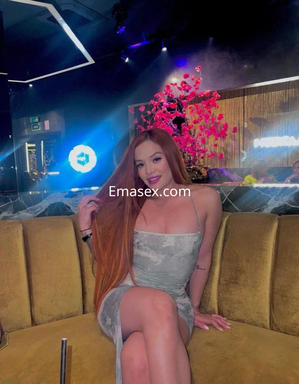 Escort en Alicante, sentada en un sofá amarillo, con un vestido plateado y cabello largo y rojizo, sonriendo en un ambiente elegante.