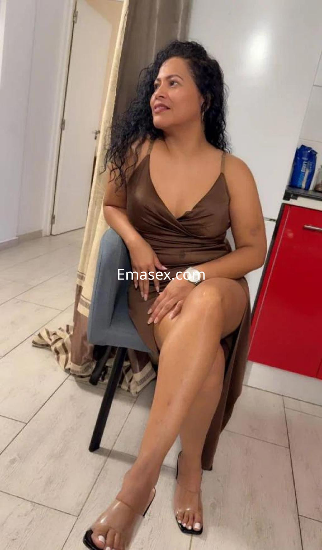 Escort en Badajoz sentada, luce un vestido marrón con tirantes y sandalias transparentes, cabello rizado.