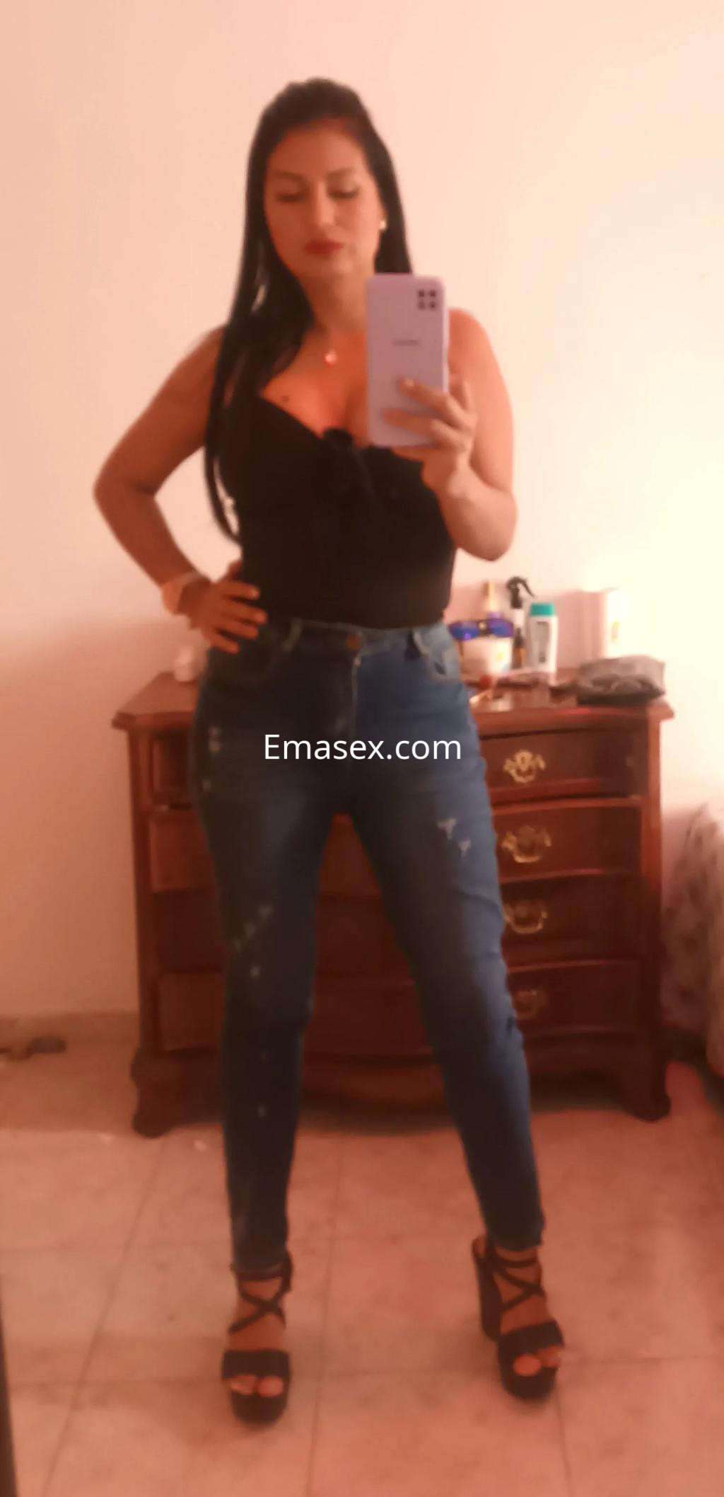 Escort en Palma de Mallorca con blusa negra y jeans ajustados, posa en un entorno interior con un espejo.