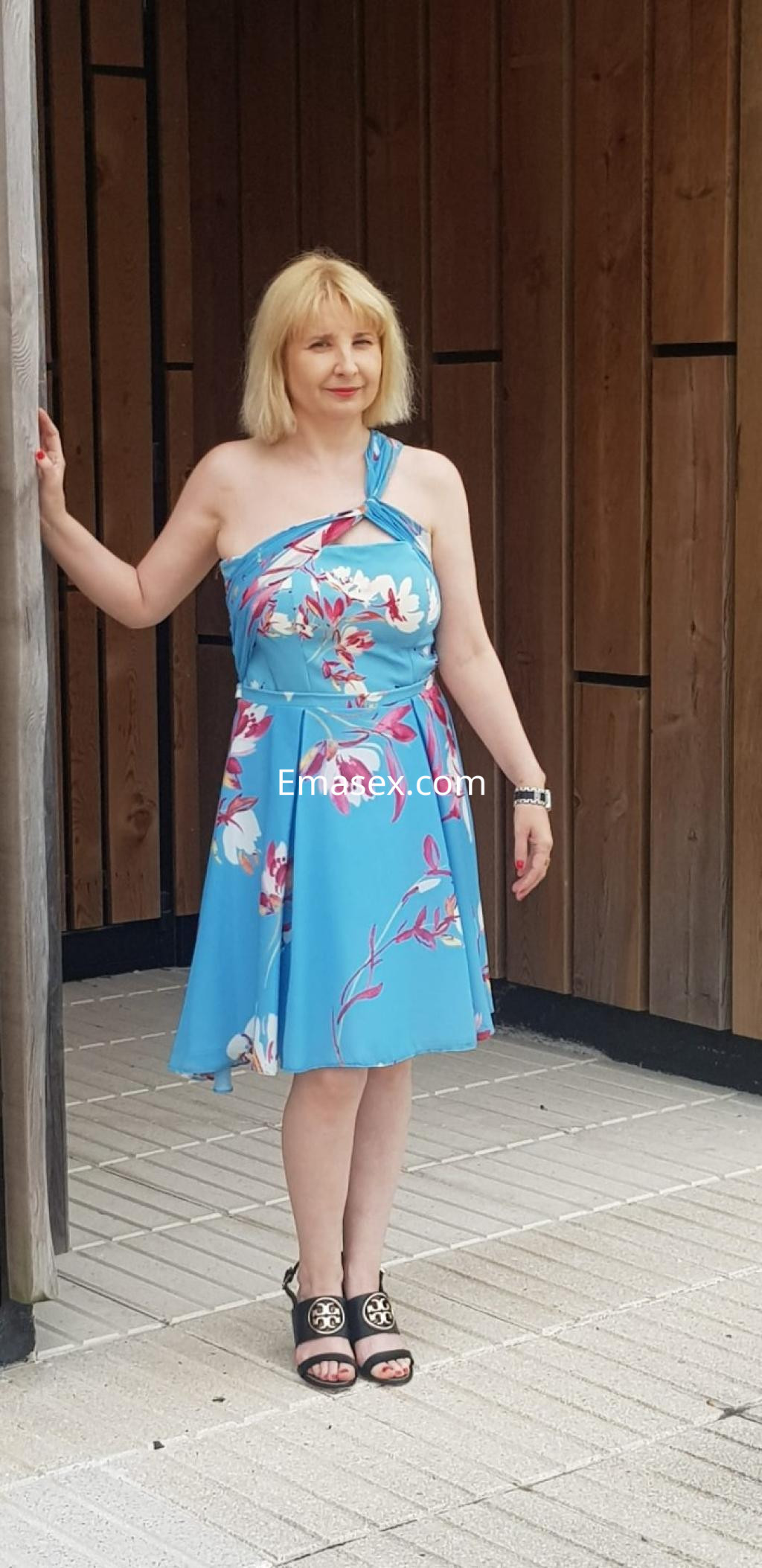 La puta viste un vestido azul con flores y sandalias de tacón, posando en un entorno moderno en Santiago.