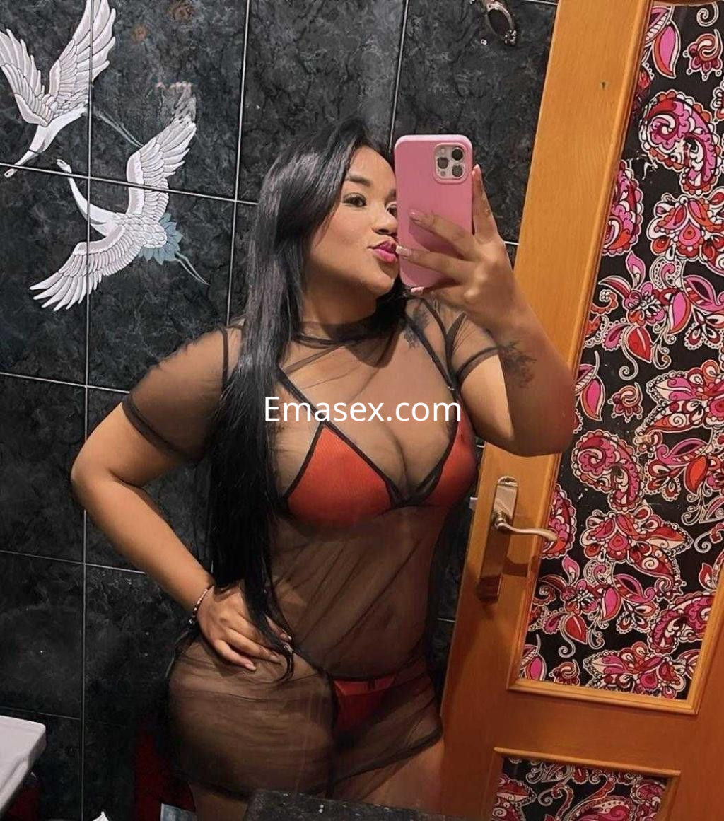 Escort en Granada posa con un conjunto de lencería roja y un vestido transparente, mostrando una expresión coqueta.
