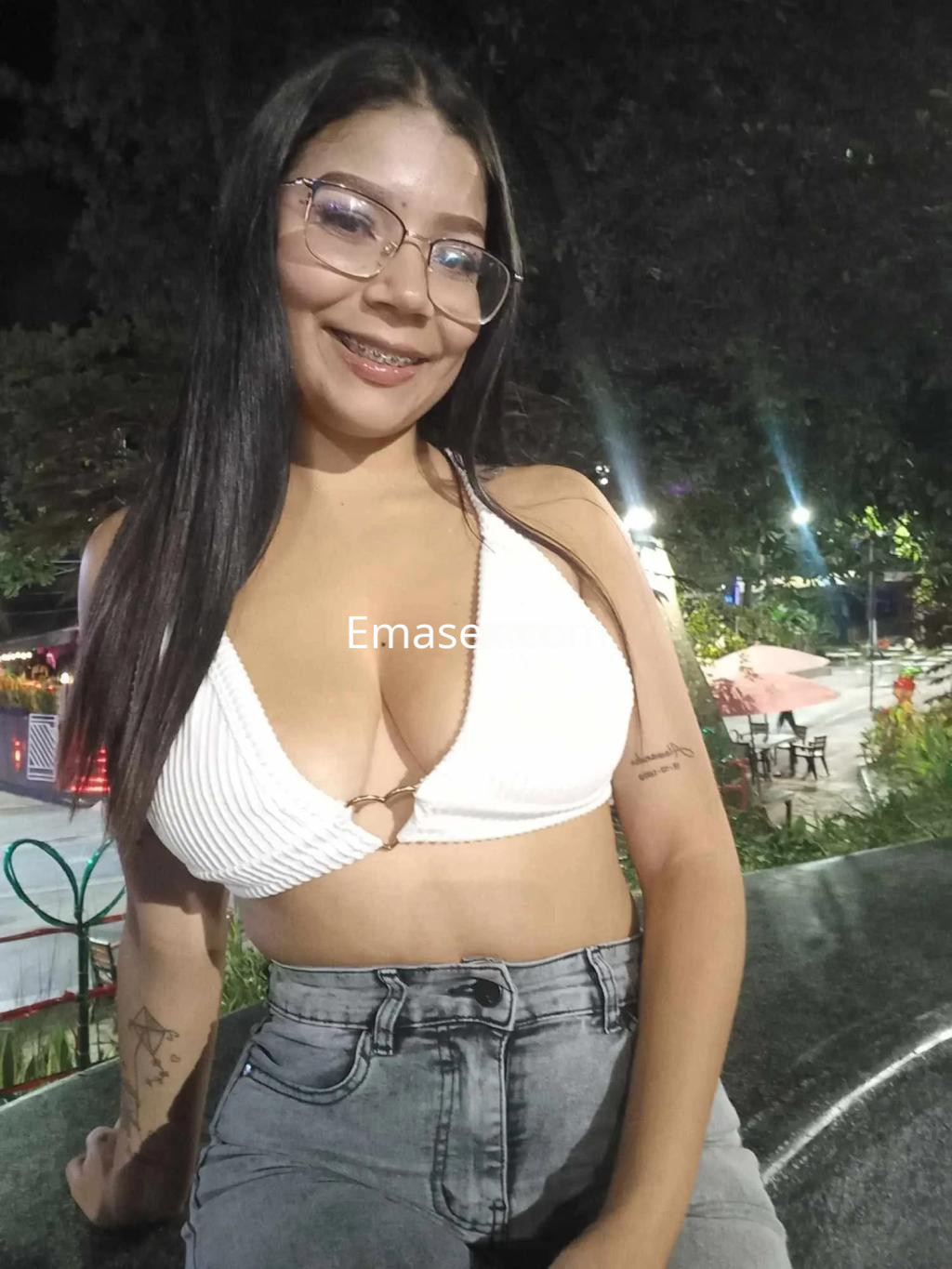 Escort en Oviedo viste un top blanco de tirantes y jeans, sonriendo con gafas en un ambiente al aire libre.